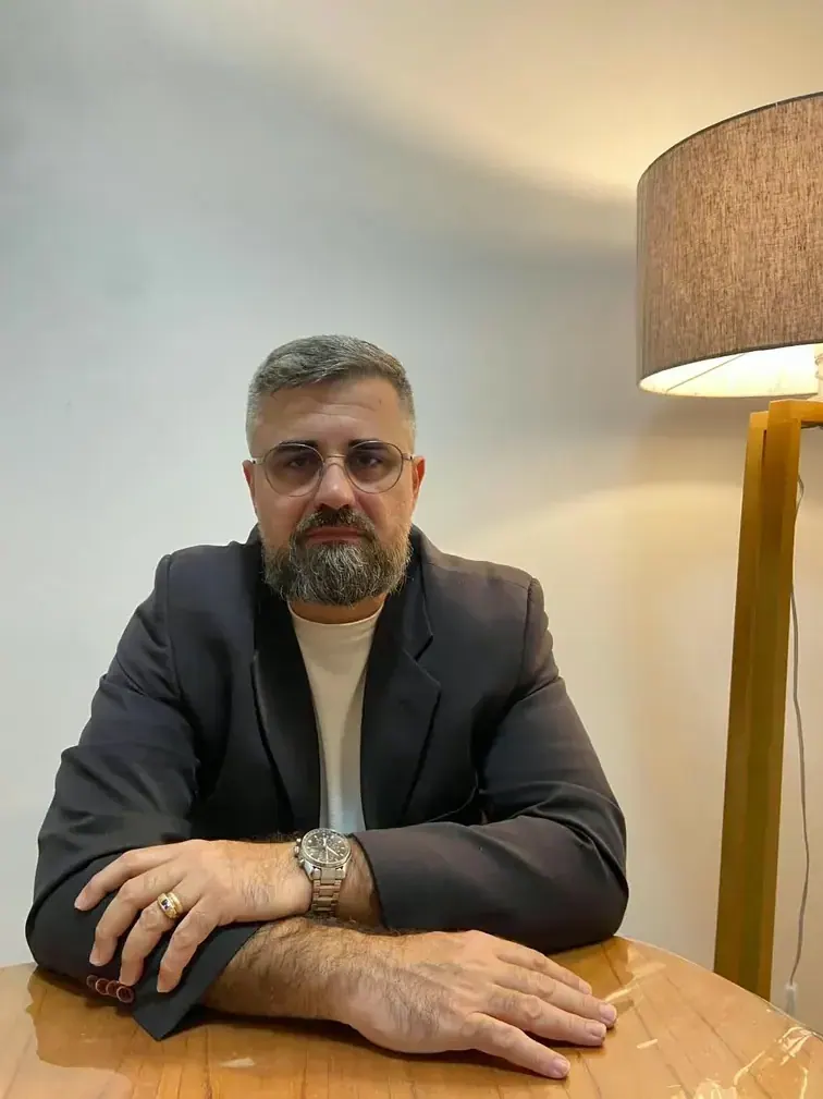 Antônio Júnior sentado à mesa de madeira do consultório, com uma luminária de palha ao fundo